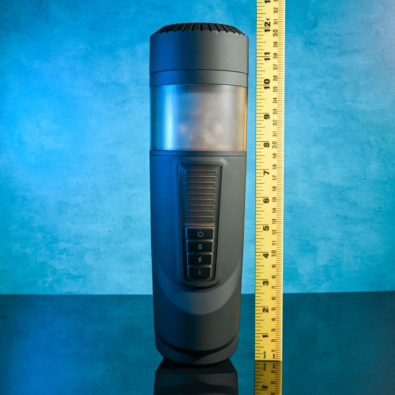 The Gender X MESSAGE IN A BOTTLE - USB Rechargeable Thrusting & Spinning Masturbator, with its turbo mode and front buttons, stands next to a yellow measuring ruler. Its reflective surface against a textured blue wall highlights the translucent section, ideal for photography or display.