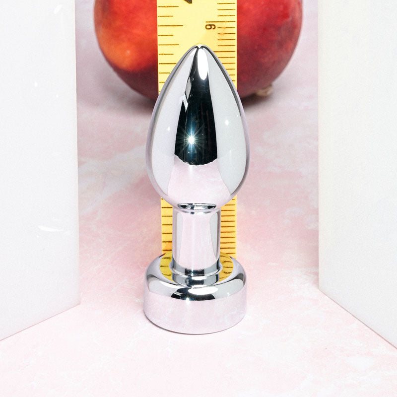 The Gender X ROCKIN METAL PLUG—a 9.3 cm metallic, USB rechargeable butt plug—stands upright between two white panels on a pink surface. A yellow ruler and a red apple are partially visible behind it. Its mirror-like finish hints at waterproof elegance and anal stimulation capability.