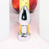 The Gender X ROCKIN METAL PLUG—a 9.3 cm metallic, USB rechargeable butt plug—stands upright between two white panels on a pink surface. A yellow ruler and a red apple are partially visible behind it. Its mirror-like finish hints at waterproof elegance and anal stimulation capability.