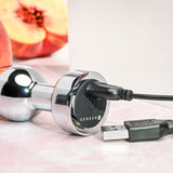 A shiny chrome Gender X ROCKIN METAL PLUG, a 9.3 cm USB-rechargeable butt plug, features prominently with its logo on the base. Nearby, a USB cable connects to another plug, while in the blurred background, a peach suggests playful waterproof fun.