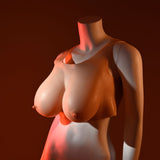 A mannequin torso displays the beige silicone Gender X UNDERGARMENTS - PLATE E-CUP, Light - Flesh Wearable Breasts, E-Cup Size. Warm lighting casts dramatic shadows against the dark backdrop, accentuating its curves and contours.