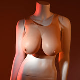 Dramatic red and orange lighting highlight the curves of a female mannequin torso, showcasing Gender X UNDERGARMENTS - PLATE E-CUP - Light silicone vest enhancements in contrast with its smooth, pale surface against a rich, dark brown background.