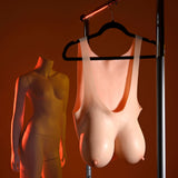 A headless mannequin with a female torso stands beside a set of Gender X UNDERGARMENTS - PLATE E-CUP - Light, realistic flesh wearable breasts, hanging on a black hanger. The scenes warm orange glow casts shadows, highlighting their lifelike texture and appearance.