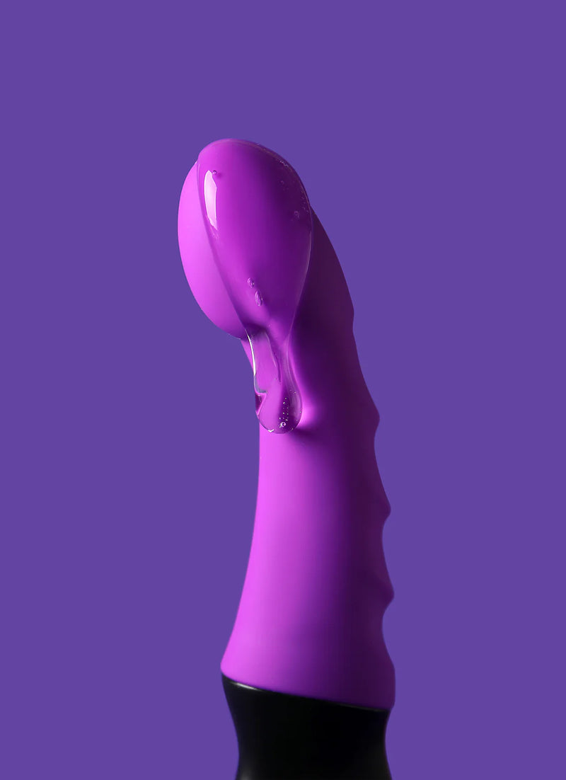 Purple silicone object against a purple background. The object has a curved, bulbous tip and a textured, ribbed body. A glossy, translucent substance drips over the top.