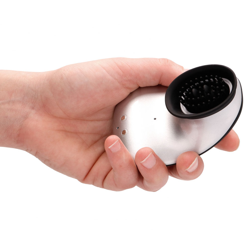 A hand holds the Twitch 1 Silver USB Rechargeable Suction Vibrator, an oval-shaped silver device for clitoral stimulation featuring a textured black circular opening on top and two button-like features. The plain white background highlights the intimate product and the gentle grip of the hand.