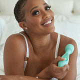 A smiling person with short hair holds the JimmyJane Canna - Teal 21.8 cm Massage Wand, known for its powerful vibrations. They wear a white top and gold earrings, sitting on a bed with white sheets in a bright, clean setting that creates a relaxed and comfortable mood.