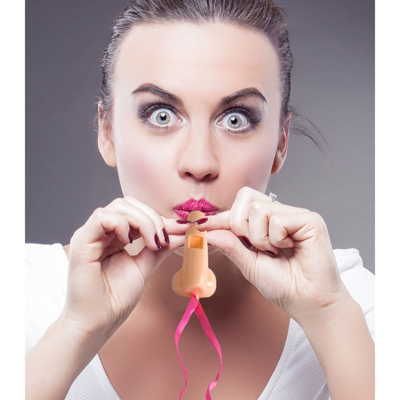 A woman with wide eyes and red lipstick playfully holds a Super Fun Penis Party Whistle to her lips, her dark hair pulled back, complementing her white top. The novelty whistle has pink streamers attached, adding flair against the gray background.