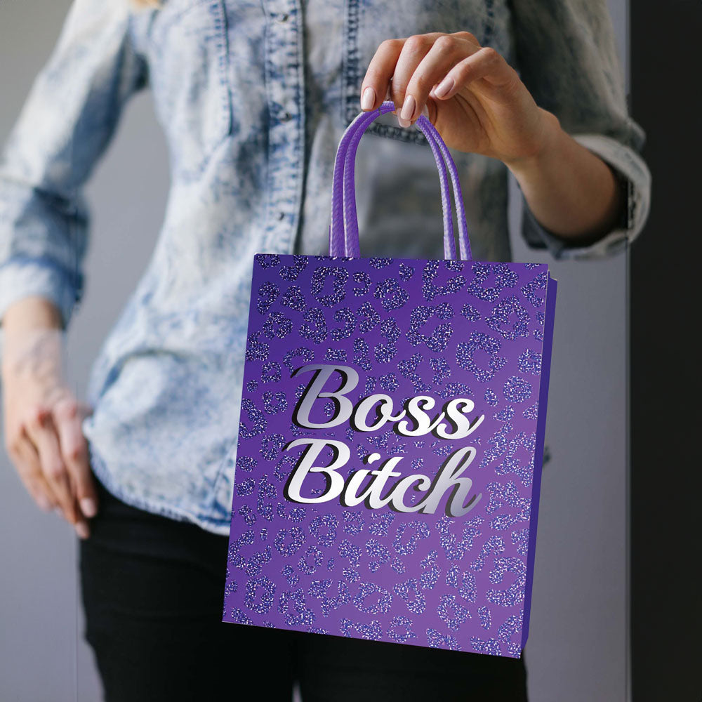 A person in a denim shirt holds a purple Boss Bitch gift bag made from strong card stock, featuring an elegant woven handle and a small blue and white pattern. Their left hand rests on their hip, showing only part of their torso and arm.