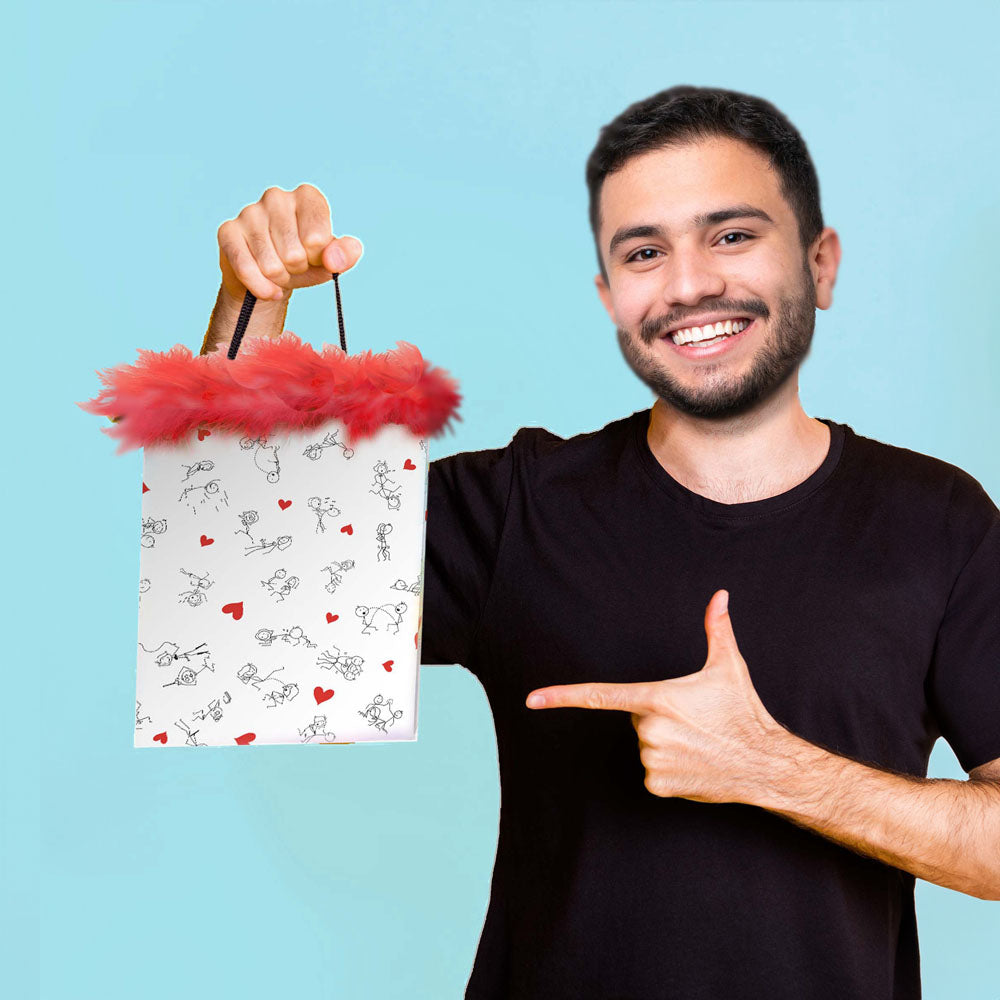 A smiling person in a black t-shirt holds up the Naughty Stick Figures Gift Bag - Novelty Gift Bag with one hand and points to it with the other. The white bag showcases playful, dirty stick figures and red hearts, trimmed with red feathers. The background is solid light blue.