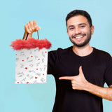 A smiling person in a black t-shirt holds up the Naughty Stick Figures Gift Bag - Novelty Gift Bag with one hand and points to it with the other. The white bag showcases playful, dirty stick figures and red hearts, trimmed with red feathers. The background is solid light blue.