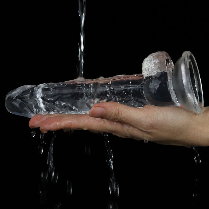 A hand holds the Flawless Clear Dildo 7.5 - Clear 19 cm Dong under running water against a black background, highlighting its skin-safe silicone and transparent texture, as glistening water droplets cascade over its phallic shape, creating a shiny, wet appearance.