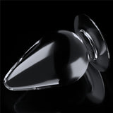 The Flawless Clear Anal Plug 4.5 - Clear 11.5 cm Butt Plug, teardrop-shaped with a flat circular base, sits on a reflective black surface. Its smooth glass design catches the light, creating subtle highlights and soft reflections beneath.