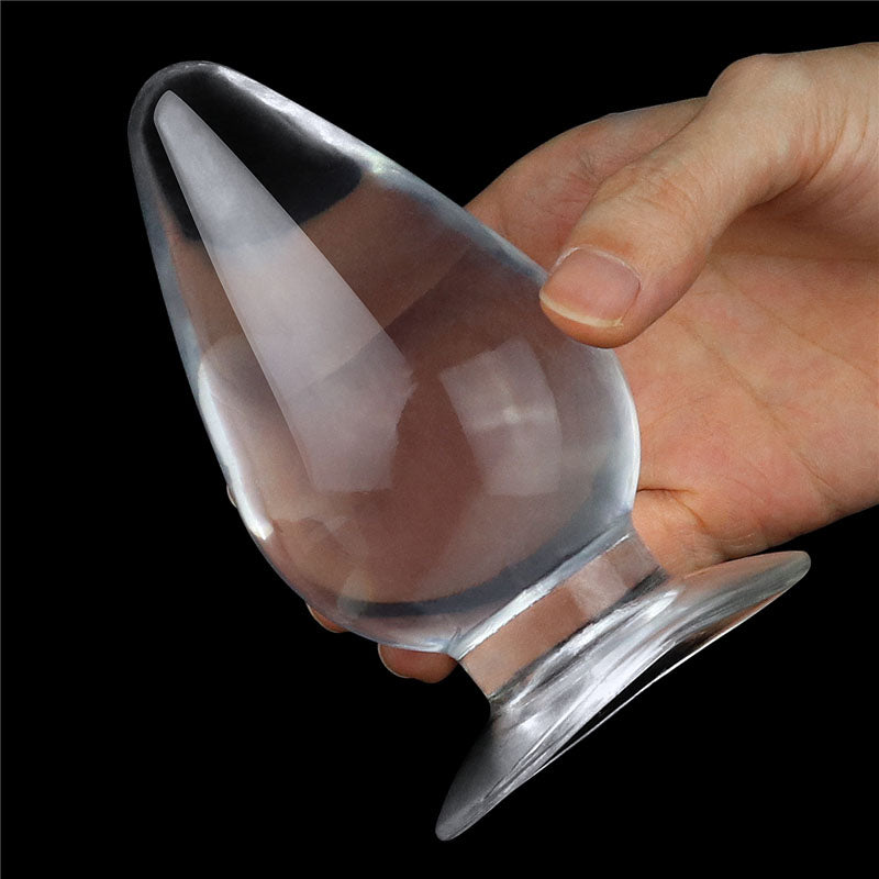 A hand holds the Flawless Clear Anal Plug 4.5, a teardrop-shaped, sculptural glass object with a flared base against a black background. This smooth, skin-safe butt plug reflects light and highlights its translucent areas.
.