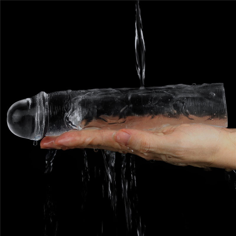 A hand holds the Flawless Clear Penis Sleeve 2 under running water against a black background, with water cascading over it. The smooth, reflective sleeve has light highlighting its transparent contours.