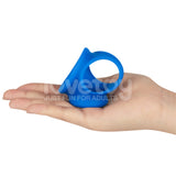 A hand holds the Pinnacle Pro Trio Grip Enhancer, a blue silicone ring with an ergonomic design. It features a circular loop at the top and a flared base. The words love toy JUST FUN FOR ADULTS appear faintly against the plain white background.