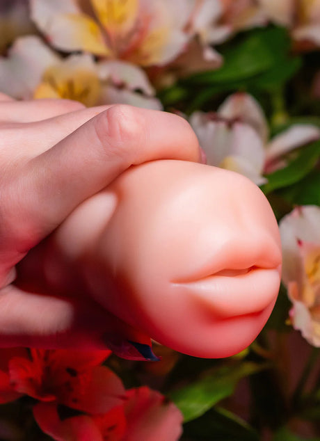 A hand is squeezing a small, artificial object resembling a pair of lips against a background of vibrant, partially blurred flowers. The lighting is soft, highlighting the smooth texture of the lips and the vivid colors of the blossoms.