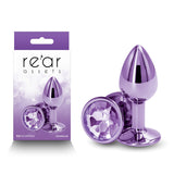 The image shows the Rear Assets Small Butt Plug in purple aluminum with a round gem base, next to its packaging. This stylish and body-safe anal toy features an enchanting design, highlighted by its jewel-like detail.