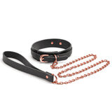 The Bondage Couture Collar & Leash - Black Restraint set, featuring rose gold metallic buckles and chain, lies elegantly displayed. The synthetic leash has a looped handle and connects to the matching black leather collar for a luxurious touch.