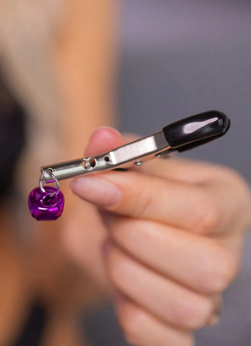 Close-up of a hand holding a metallic clip with a black handle. Attached to the clip is a small, shiny purple bell hanging from a circular loop. The background is blurred, highlighting the clip and bell in focus.