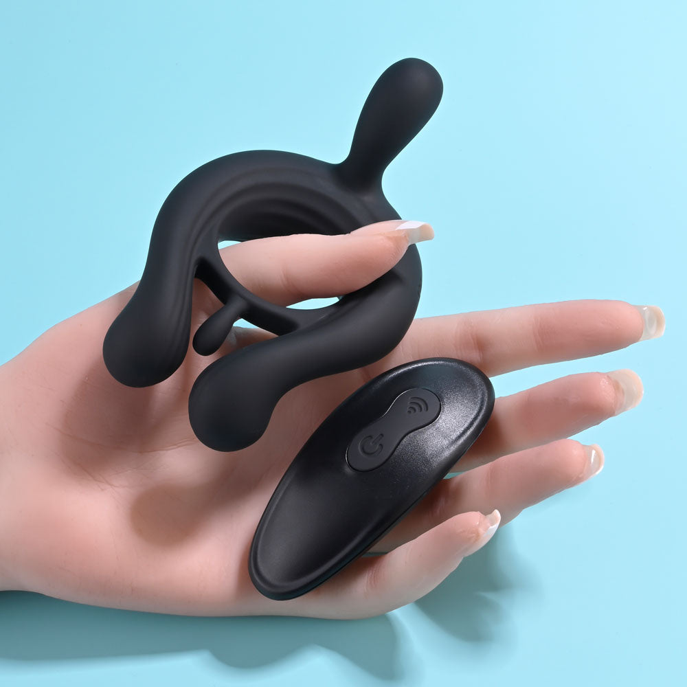 The Playboy Pleasure TRIPLE PLAY, a sleek black silicone cock ring with curved extensions, rests on a hand with manicured nails. Paired with an oval remote control featuring two buttons on a light blue background, it offers robust vibrating motors and wireless functionality.