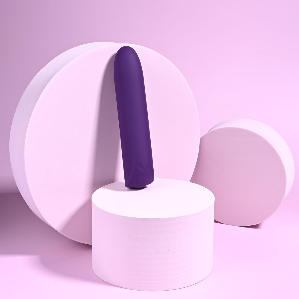 The Playboy Pleasure ONE & ONLY, a purple 12 cm USB rechargeable bullet vibrator made of waterproof silicone, stands gracefully on a white pedestal. Its minimalist design complements the larger circular structures and pastel pink backdrop, with left-casting shadows adding depth.