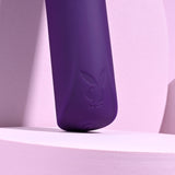 A close-up of the Playboy Pleasure ONE & ONLY - Purple 12 cm USB Rechargeable Bullet reveals its waterproof silicone design with a subtle embossed logo of a bunny in a bowtie. The bullet rests on curved, light pink surfaces casting soft shadows for a sleek, modern look.