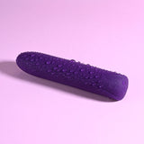 A purple Playboy Pleasure ONE & ONLY bullet vibrator rests on a pastel pink surface. Water droplets add gloss to the waterproof toy, highlighting its smooth, gently tapered shape.