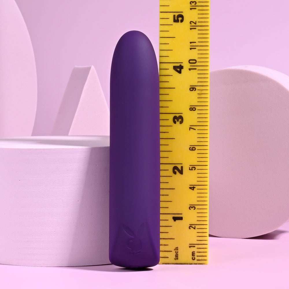 The Playboy Pleasure ONE & ONLY - Purple 12 cm USB Rechargeable Bullet, a purple cylindrical silicone device, stands upright against a yellow inch-marked measuring tape on a light pink surface with pastel pink abstract geometric shapes in the background.