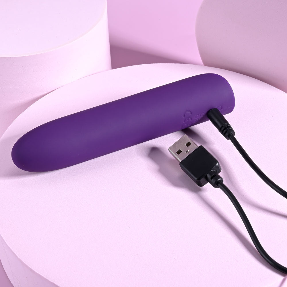 The Playboy Pleasure ONE & ONLY - Purple 12 cm USB Rechargeable Bullet, crafted from silicone, rests on a pastel pink surface. Its linked to a black USB charging cable, with rounded pastel pink shapes in the background creating a soothing atmosphere.