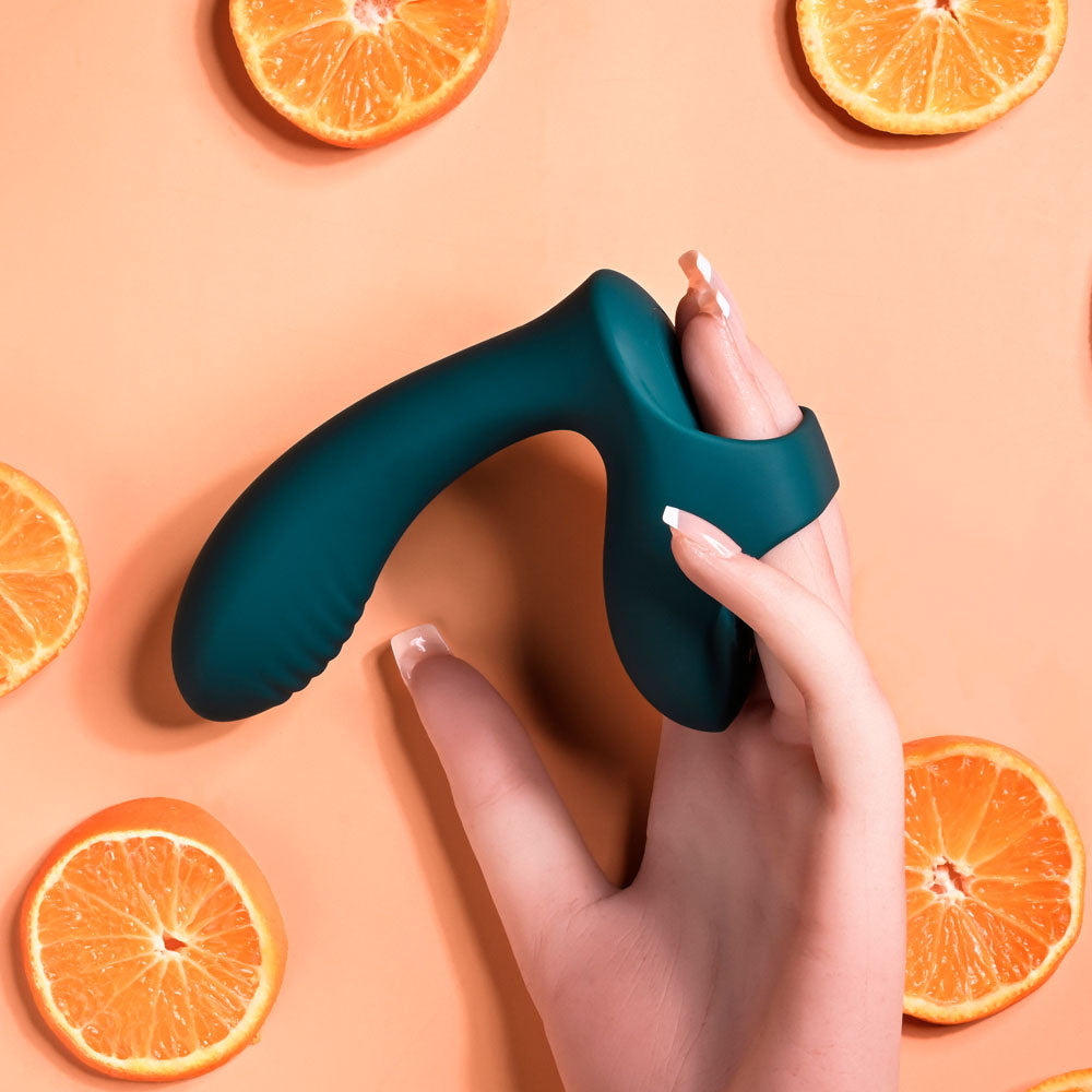 A hand holds the green Playboy Pleasure WRAPPED AROUND YOUR FINGER USB Rechargeable Finger Stimulator, characterized by a curved shape and loop handle, against a peach backdrop. Cut orange halves surround it, while the persons nails, manicured with white tips, add elegance to the scene.