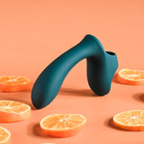 A green silicone Playboy Pleasure WRAPPED AROUND YOUR FINGER stimulator stands upright on a peach surface, surrounded by halved oranges. This ergonomic finger vibrator offers G-Spot and P-Spot stimulation with its curved design and comfortable grip, creating an inviting, playful atmosphere.