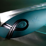 The Playboy Pleasure PINPOINT PERFECTION is a sleek, green 19.3 cm vibrator made from body-safe silicone with a black cross design and power button. It boasts an embossed rabbit logo, ergonomic shape, and powerful vibrations highlighted under soft lighting on a reflective surface.