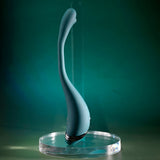 The Playboy Pleasure PINPOINT PERFECTION, a USB rechargeable vibrator standing upright against a dark green background, showcases its teal, ergonomic design with a smooth surface and curved shape. Metallic accents near the base add an elegant touch to the 19.3 cm poseable device.