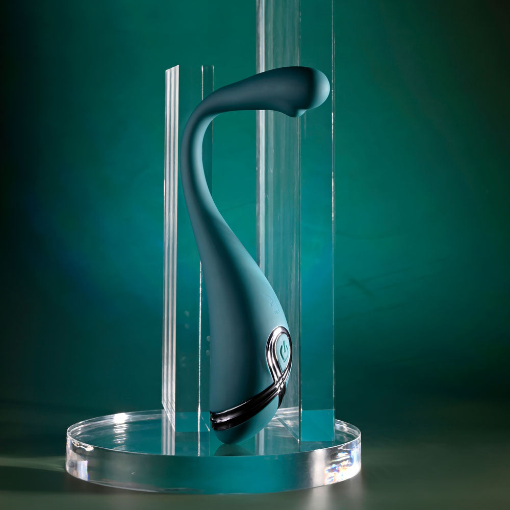 The Playboy Pleasure PINPOINT PERFECTION is a sleek, green 19.3 cm USB rechargeable vibrator. Made from body-safe silicone with powerful vibrations, it features a bulbous tip and clear vertical panels that emphasize its design on a transparent stand against a gradient of dark to light teal.