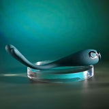 The Playboy Pleasure PINPOINT PERFECTION vibrator, a dark green beauty crafted from body-safe silicone with a bulbous end and loop handle, is elegantly displayed on a clear acrylic stand. The teal and dark green backdrop highlights its powerful vibrations within the streamlined design.