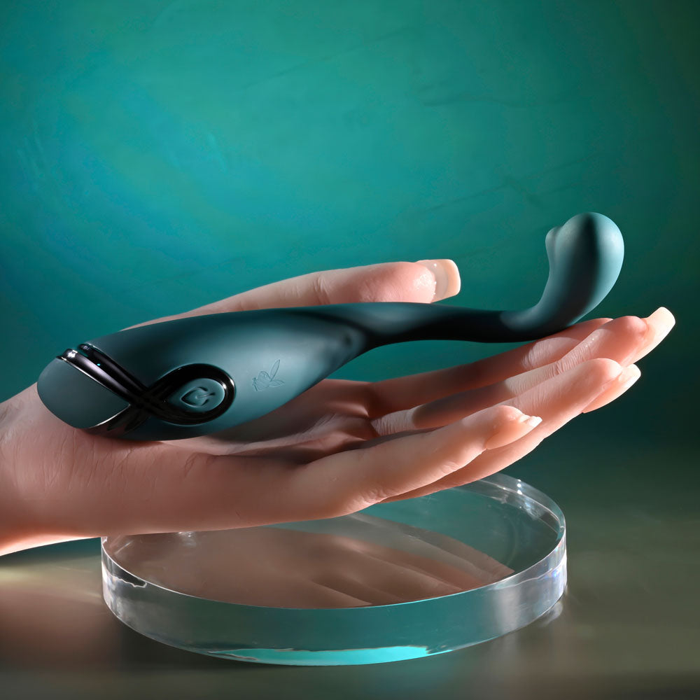 A green Playboy Pleasure PINPOINT PERFECTION 19.3 cm poseable vibrator, crafted from body-safe silicone, rests in a realistic sculpted hand against a teal gradient. The hand is on a transparent circular platform, adding an artistic and futuristic touch to the image.