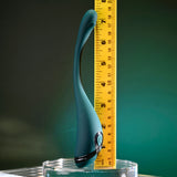 A sleek, teal Playboy Pleasure PINPOINT PERFECTION vibrator made from body-safe silicone stands upright on a glass surface beside a partially retracted yellow measuring tape. The background features a modern gradient from dark green to teal.