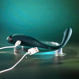 The Playboy Pleasure PINPOINT PERFECTION Vibrator, in sleek dark green and crafted from body-safe silicone, features a curved design on a clear stand. It connects to a white USB cable, set against a modern green gradient backdrop for an elegant touch.