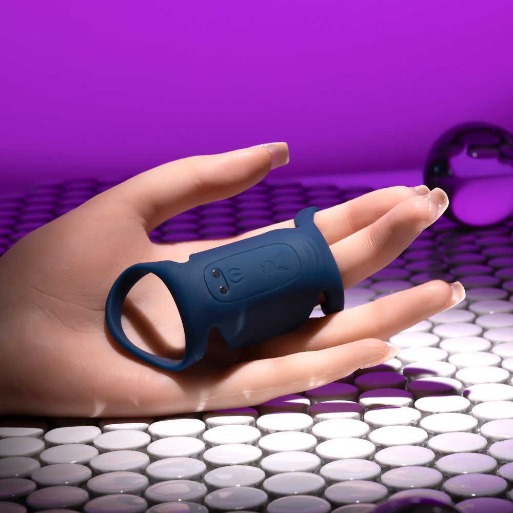 A realistic model hand on a reflective, tiled surface holds the Playboy Pleasure JET PACK in dark blue, a body-safe silicone vibrating penis harness, around the index and middle fingers. It features small buttons and is waterproof. The background is a purple gradient with a reflective sphere on the right.