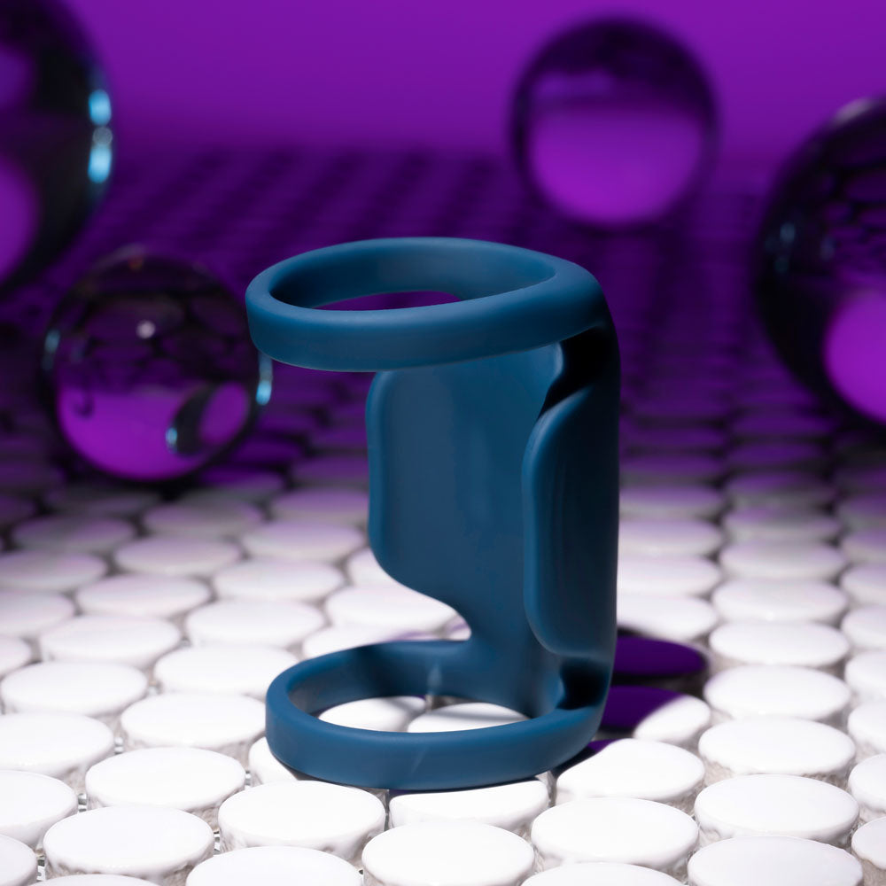 A modern ambiance is created by the Playboy Pleasure JET PACK - Dark Blue USB Rechargeable Vibrating Penis Harness, whose abstract sculpture of circular elements, made from body-safe silicone, stands on white round tiles with translucent purple spheres and a purple-to-black gradient backdrop.