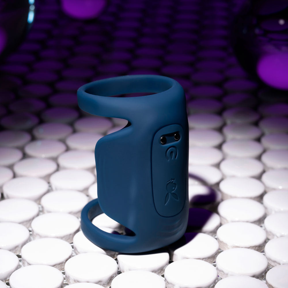 A dark blue Playboy Pleasure JET PACK penis vibrator, crafted from body-safe silicone with two black side buttons, rests on a reflective white tile. Blurred purple and black hues in the background create a futuristic atmosphere.