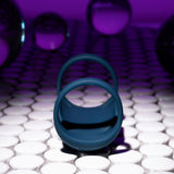 The dark blue Playboy Pleasure JET PACK, a USB rechargeable vibrating penis harness crafted from body-safe silicone, rests on a reflective white tiled floor. Translucent spheres hover in the background as purple lighting enhances the modern and surreal ambiance.