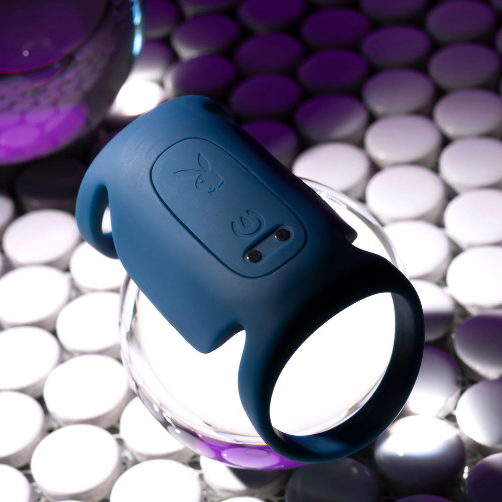 The Playboy Pleasure JET PACK - Dark Blue USB Rechargeable Vibrating Penis Harness, crafted from body-safe silicone with control buttons and an embossed logo, rests on a transparent stand. Illuminated purple-tinted circles in the background create dramatic shadows and highlight its contours.