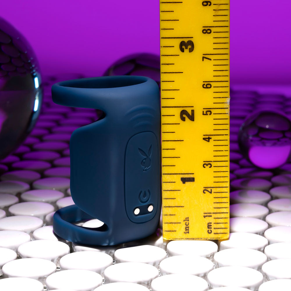 The Playboy Pleasure JET PACK, a dark blue body-safe silicone USB rechargeable vibrating penis harness with electronic components, stands beside a yellow nearly 3-inch measuring tape on a white tiled surface. The purple background enhances the view of reflective objects. It includes buttons and LED lights.