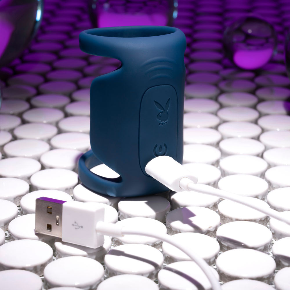 The dark blue Playboy Pleasure JET PACK, a modern silicone vibrating penis harness, is charging via a white USB cable on a glossy round-tiled surface. Surrounded by purple light reflections and glass spheres, its futuristic design is enhanced.