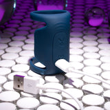 The dark blue Playboy Pleasure JET PACK, a modern silicone vibrating penis harness, is charging via a white USB cable on a glossy round-tiled surface. Surrounded by purple light reflections and glass spheres, its futuristic design is enhanced.