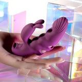 A hand holds the Playboy Pleasure LEND A HAND, a USB rechargeable purple vibrator made of body-safe silicone. It features a textured shaft and rabbit-shaped stimulator, perfect for G-spot exploration with an ergonomic design. The backdrop is iridescent holographic blocks, creating a reflective pastel environment.