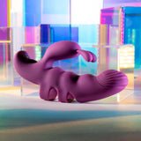 The Playboy Pleasure LEND A HAND - Purple USB Rechargeable Finger Vibrator, crafted from body-safe silicone in an animal-like form with an extended tail, sits on a reflective surface amid colorful geometric shapes. The background glows with pink, blue, and green gradients for a dreamy effect.