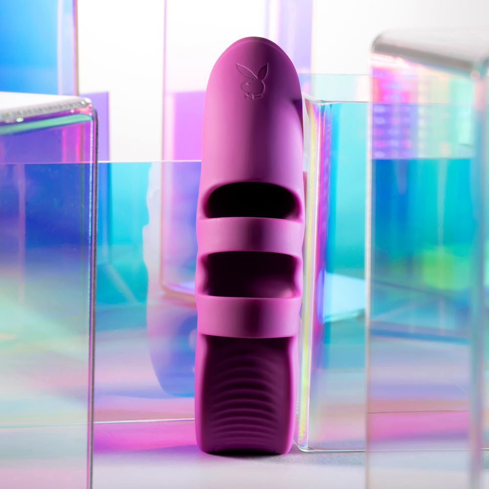 The Playboy Pleasure LEND A HAND - Purple USB Rechargeable Finger Vibrator, made of body-safe silicone with two finger loops, stands on a reflective surface amidst iridescent acrylic panels, offering a vibrant abstract background ideal for G-Spot exploration.