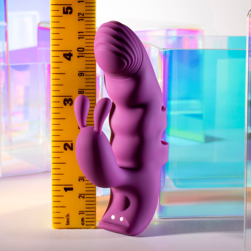 Next to a yellow ruler, the purple Playboy Pleasure LEND A HAND USB Rechargeable Finger Vibrator stands upright against a backdrop of pastel square shapes. Made from body-safe silicone, this dual motor toy is designed with textured details and a dual-pronged shape for enhanced G-spot exploration.